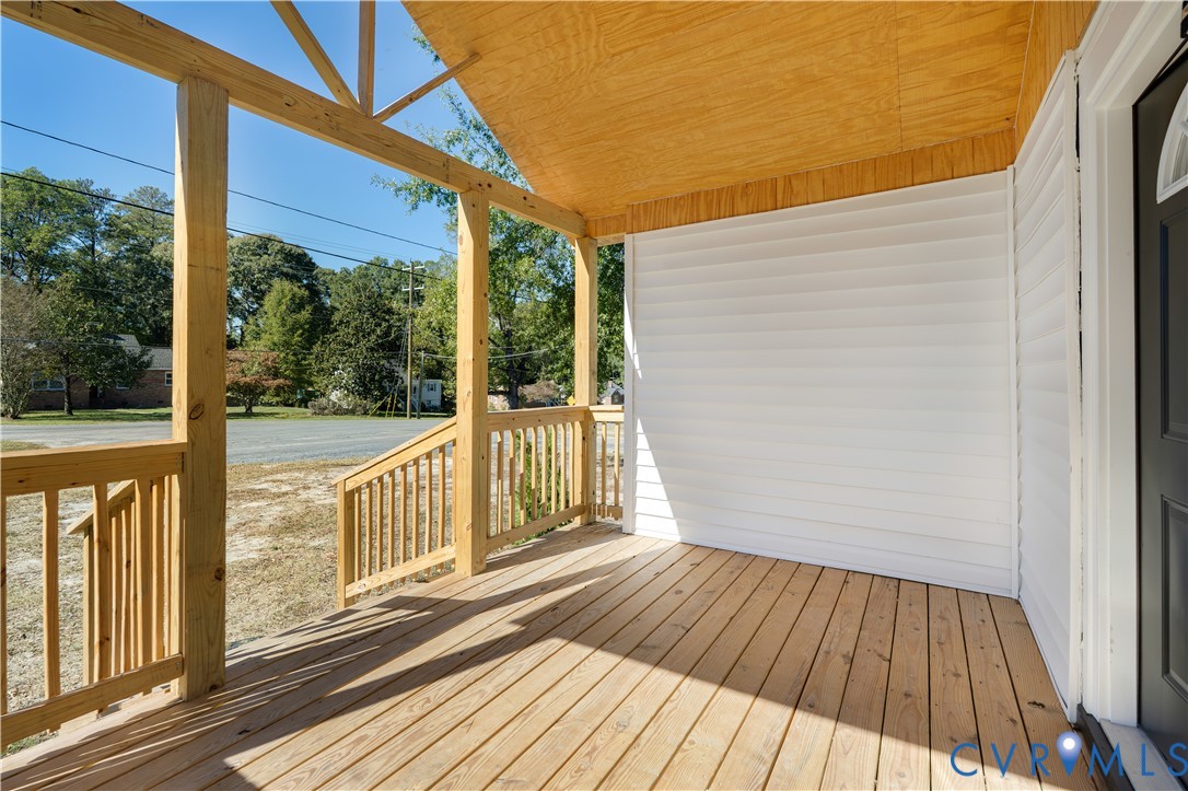 Covered front porch featuring exposed wood support beams, natural wood-finish decking, white horizontal siding, and wooden balustrade railings