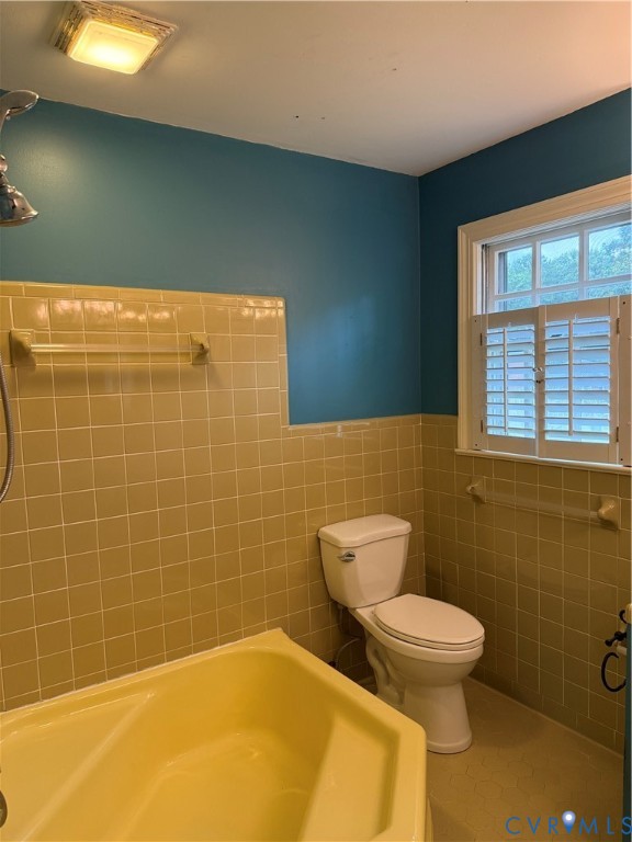 Bathroom featuring a soaking tub, ceramic tile wall surround, two-piece toilet, ceiling-mounted light fixture, and window with built-in shutters