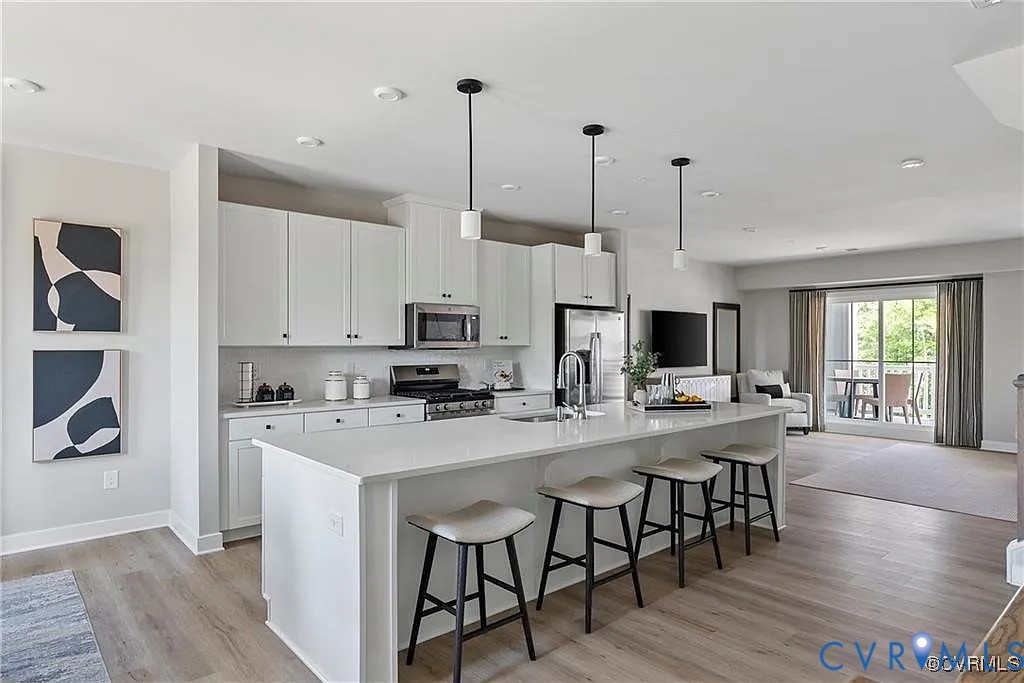 Kitchen featuring white cabinetry, an expansive central island with a solid-surface countertop, stainless steel appliances, pendant lighting, and wood-finish flooring Kitchen featuring white cabinetry, an expansive central island with a solid-surface countertop, stainless steel appliances, pendant lighting, and wood-finish flooring