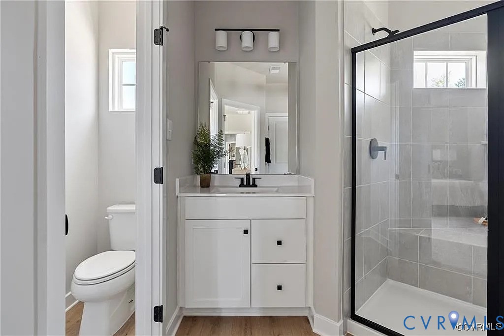 Bathroom featuring a white vanity with matte black hardware, a tiled shower with a glass enclosure, wood-finish flooring, and a three-light vanity fixture Bathroom featuring a white vanity with matte black hardware, a tiled shower with a glass enclosure, wood-finish flooring, and a three-light vanity fixture