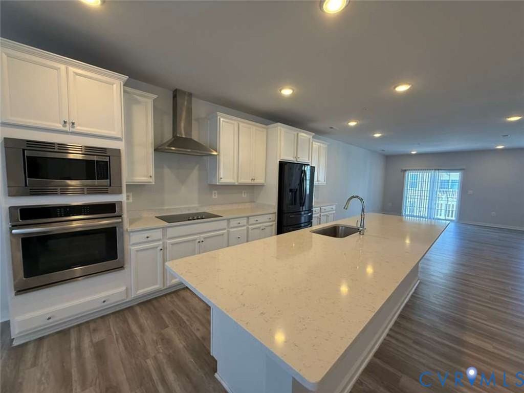 Kitchen featuring white cabinetry, quartz countertops, a center island with an integrated sink, stainless steel wall-mounted oven and microwave, and wood-finish flooring
