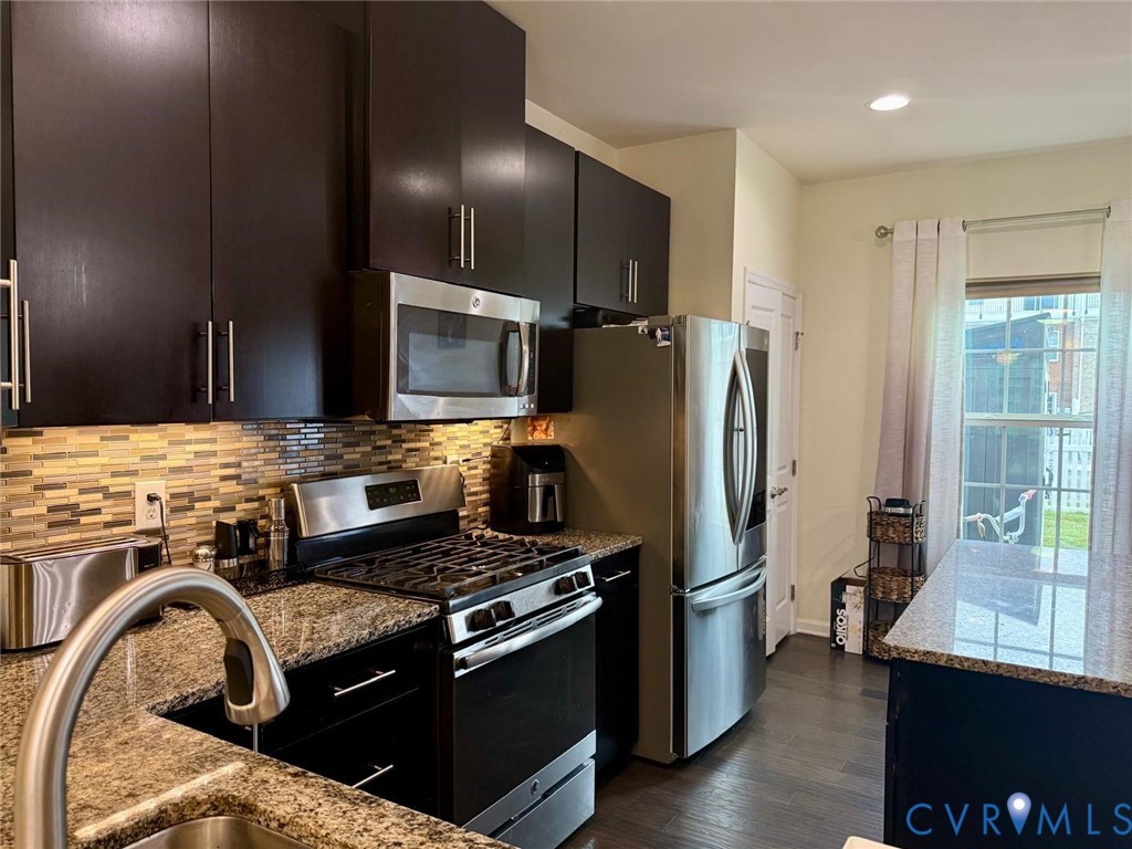 Kitchen featuring dark wood cabinetry, speckled stone countertops, a multi-tonal linear tile backsplash, stainless steel appliances, and wood-finish flooring