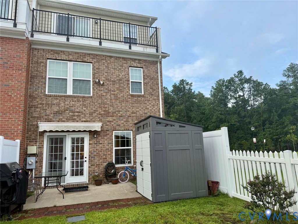 Brick exterior featuring a private concrete patio, a retractable awning, and a balcony with black metal railings, enclosed by white vinyl fencing