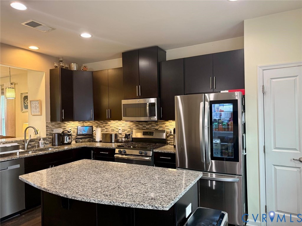 Kitchen featuring dark cabinetry, stone-surface central island, stainless steel appliances, multi-toned tile backsplash, and recessed lighting