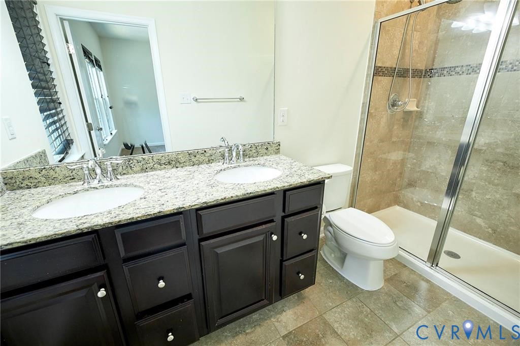 Dual-sink vanity with stone countertop and dark cabinetry, large wall-mounted mirror, and walk-in shower with stone tile surround and glass enclosure