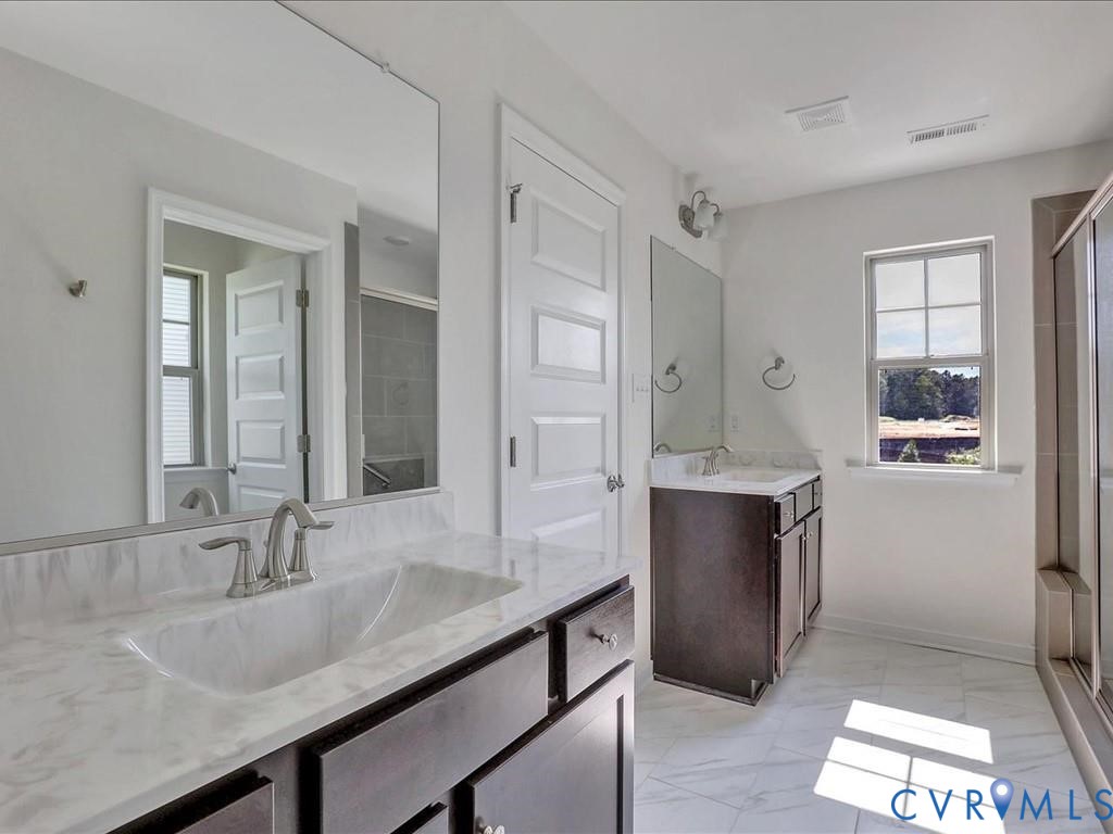 Double vanity featuring dark cabinetry, solid-surface countertops, integrated sinks with dual-handle hardware, and expansive wall-mounted mirrors Double vanity featuring dark cabinetry, solid-surface countertops, integrated sinks with dual-handle hardware, and expansive wall-mounted mirrors