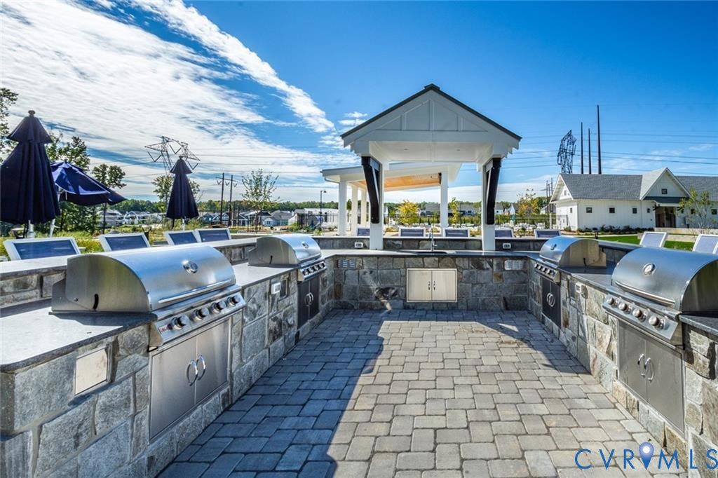 Outdoor kitchen featuring stone masonry, stainless steel built-in grills, stone paver flooring, dark-finish countertop surfaces, and an overhead pergola structure