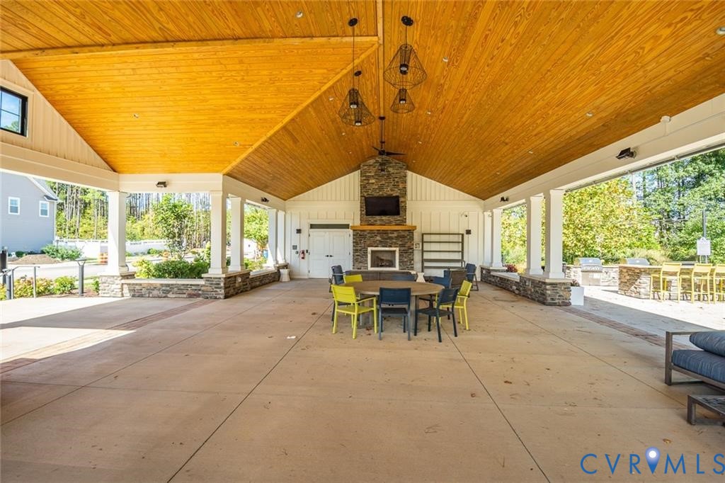 Covered outdoor pavilion featuring a vaulted wood-plank ceiling, stone-faced fireplace with wood mantel, concrete flooring, and built-in stone masonry seating