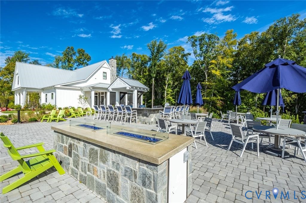 Sprawling stone paver patio featuring a built-in outdoor kitchen with stone veneer base and concrete countertop, integrated linear fire feature, and farmhouse-style exterior architecture