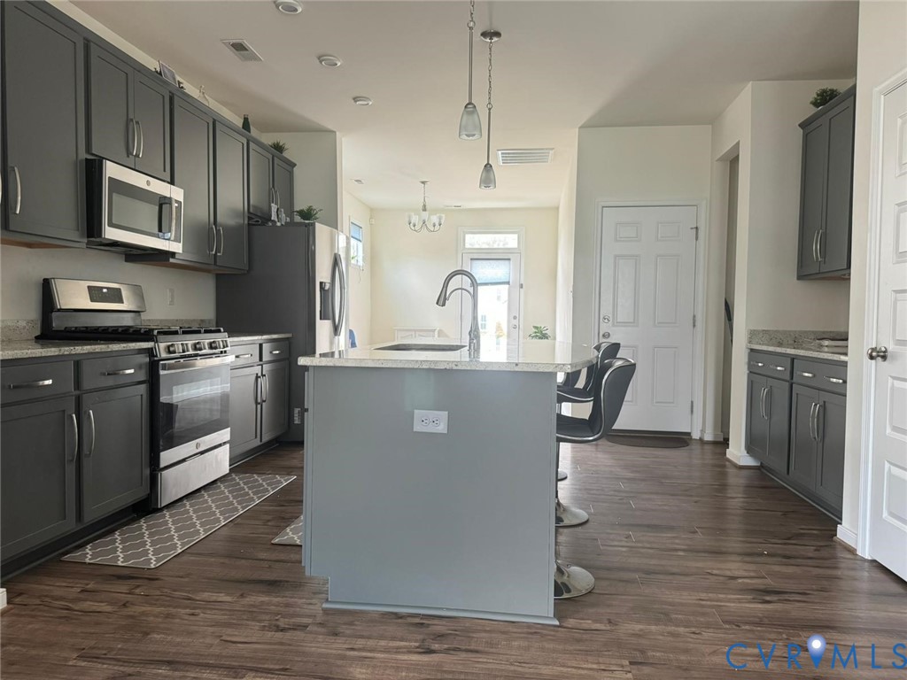 Kitchen featuring dark cabinetry, stone-finish countertops, stainless steel appliances, a central island with an integrated sink, and wood-finish flooring Kitchen featuring dark cabinetry, stone-finish countertops, stainless steel appliances, a central island with an integrated sink, and wood-finish flooring