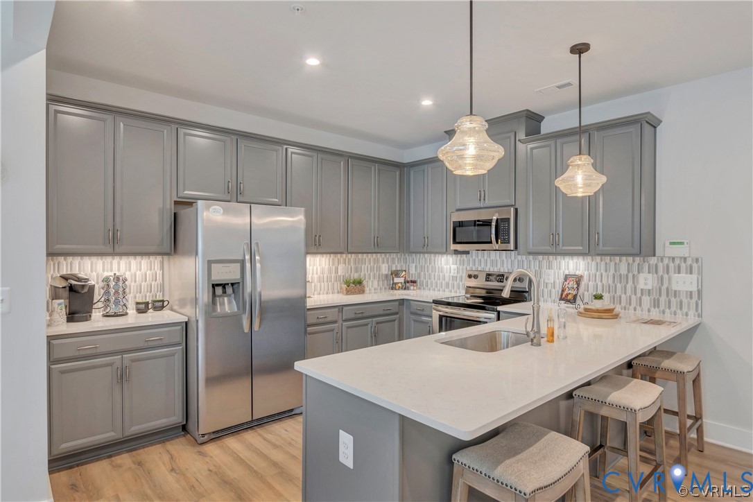 Kitchen featuring shaker-style grey cabinetry, quartz countertops, a stone-patterned tile backsplash, stainless steel appliances, and pendant lighting