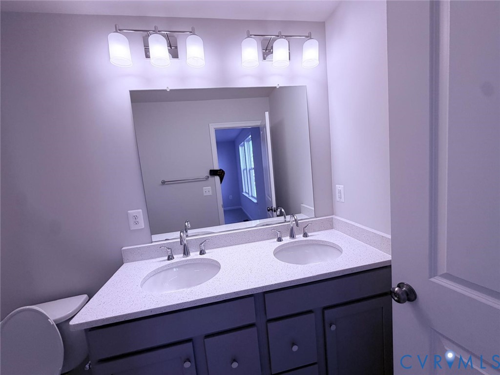 Dual-sink vanity with solid-surface countertop, cabinetry with brushed-metal hardware, large wall-mounted mirror, and multi-bulb polished-chrome vanity lighting