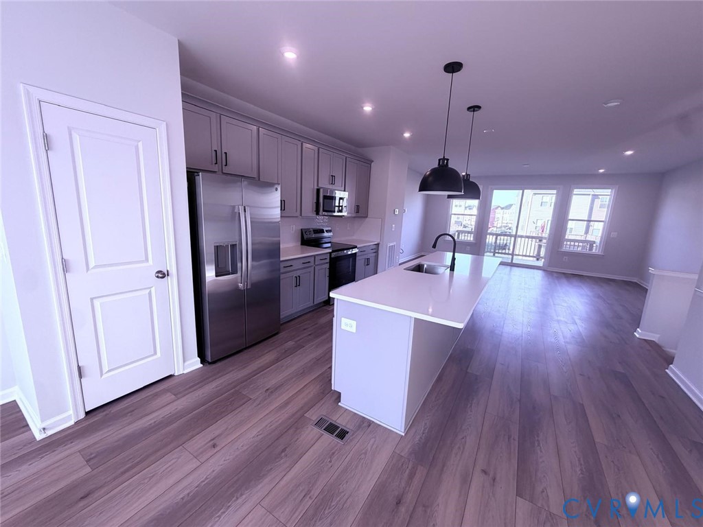 Kitchen featuring shaker-style cabinetry, quartz countertops, a central island with an integrated sink, stainless steel appliances, and wood-finish flooring