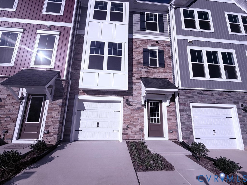 Multi-story townhouse exterior featuring mixed-material facade of stone veneer and vertical siding, covered entryways, attached garage with carriage-style doors, and multi-pane windows with shutters