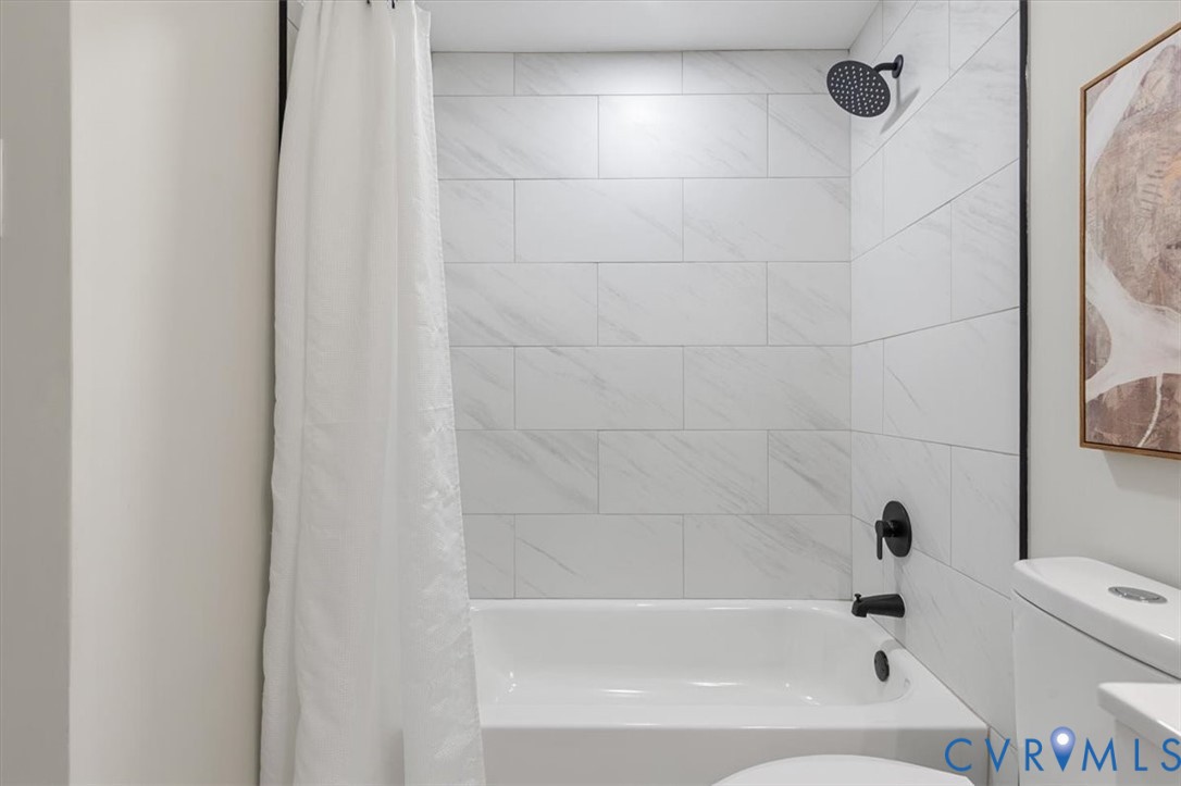 Full-height shower surround featuring large-format rectangular white tile with subtle veining, matte black rain-style shower head, matching matte black faucet and diverter, and a white soaking tub