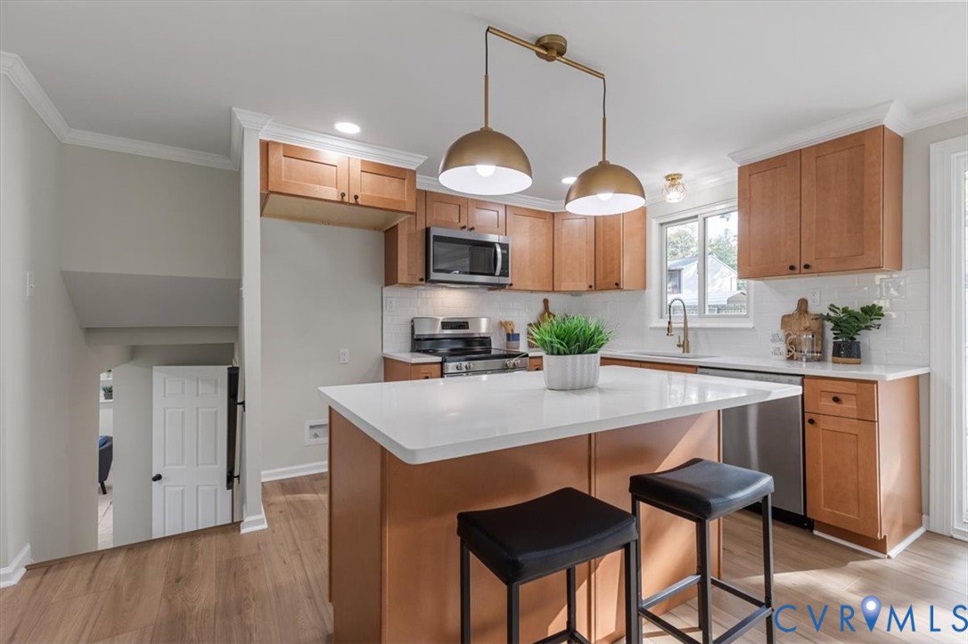 Kitchen featuring light wood-finish cabinetry, a white countertop island, stainless steel appliances, a white subway tile backsplash, and dual-pendant brass lighting
