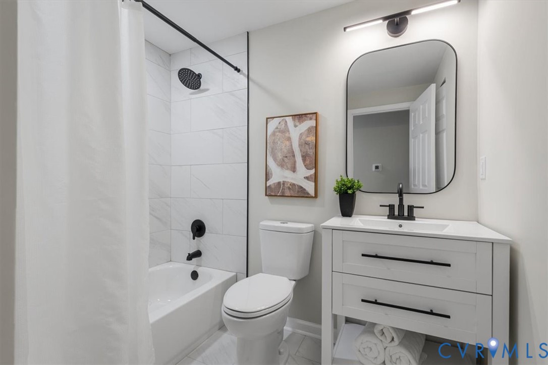 Bathroom featuring a white vanity with integrated rectangular sink, matte black hardware, stone-finish shower surround with rainfall showerhead, and a rectangular wall mirror with rounded corners