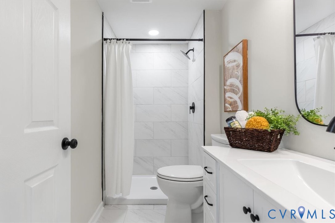 Bathroom featuring a white vanity cabinet with a stone-top integrated sink, matte black hardware, a tiled walk-in shower with black metal trim, and large-format tile flooring