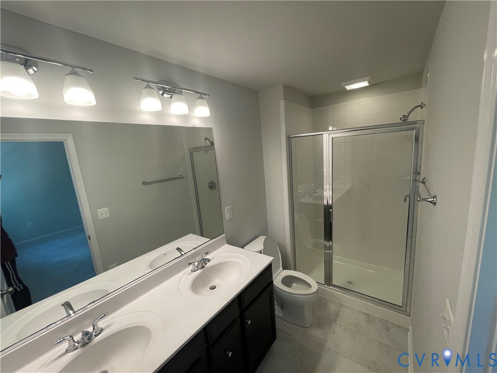 Dual-sink vanity with white countertops, dark cabinetry, full-width mirror, glass-enclosed shower, and large-format tile flooring