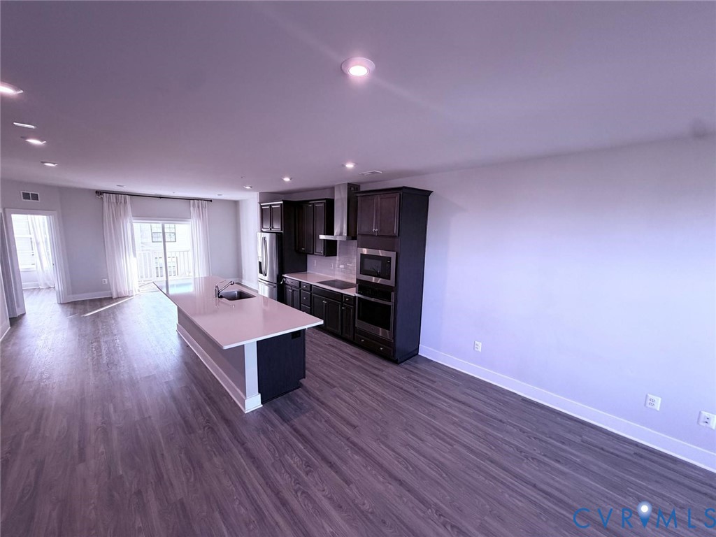 Kitchen featuring dark cabinetry, expansive stone-surface island with integrated sink, stainless steel built-in double wall oven, and wood-finish flooring throughout