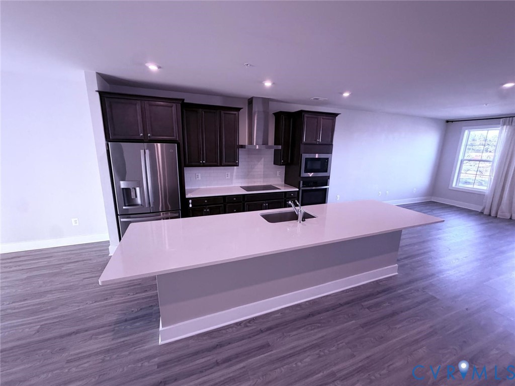 Kitchen featuring dark-finish cabinetry, stone-surface countertops, stainless steel appliances, a center island with an undermount sink, and wood-finish flooring