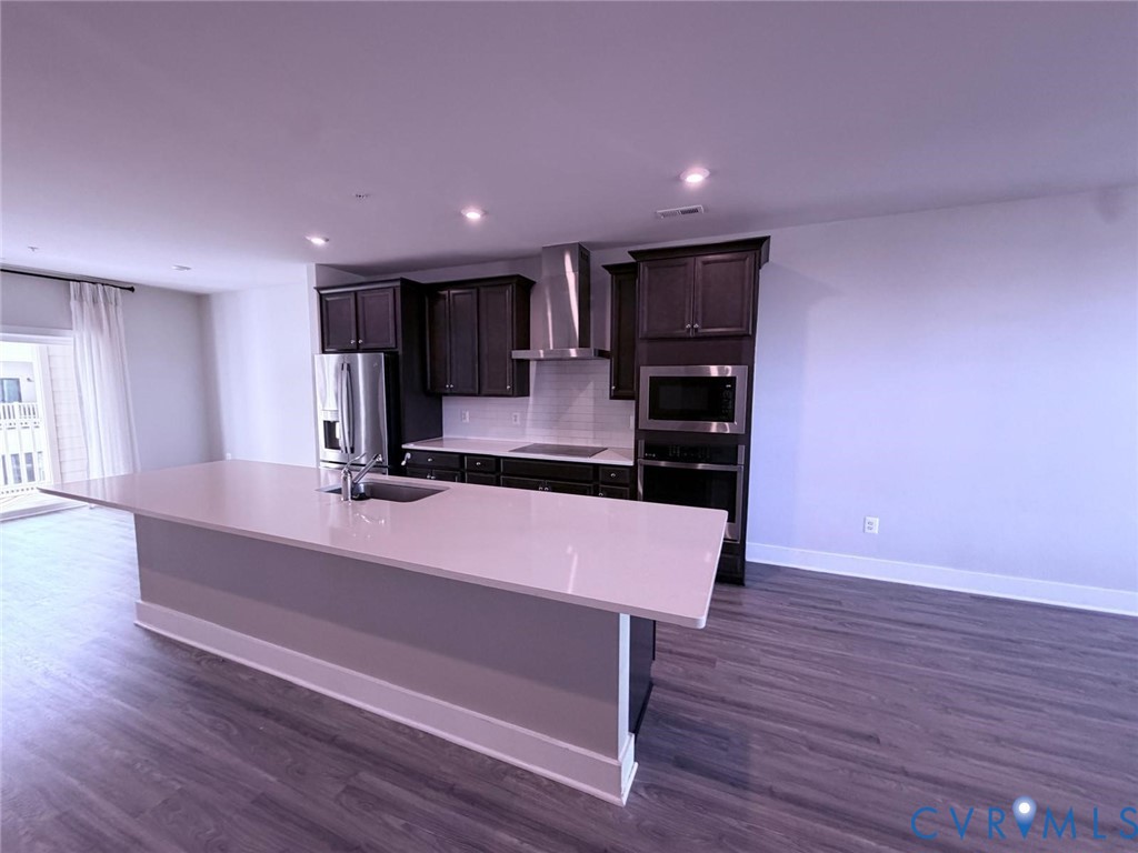 Kitchen featuring a central island with a solid surface countertop and undermount sink, dark-toned cabinetry, stainless steel appliances, recessed lighting, and wood-finish flooring