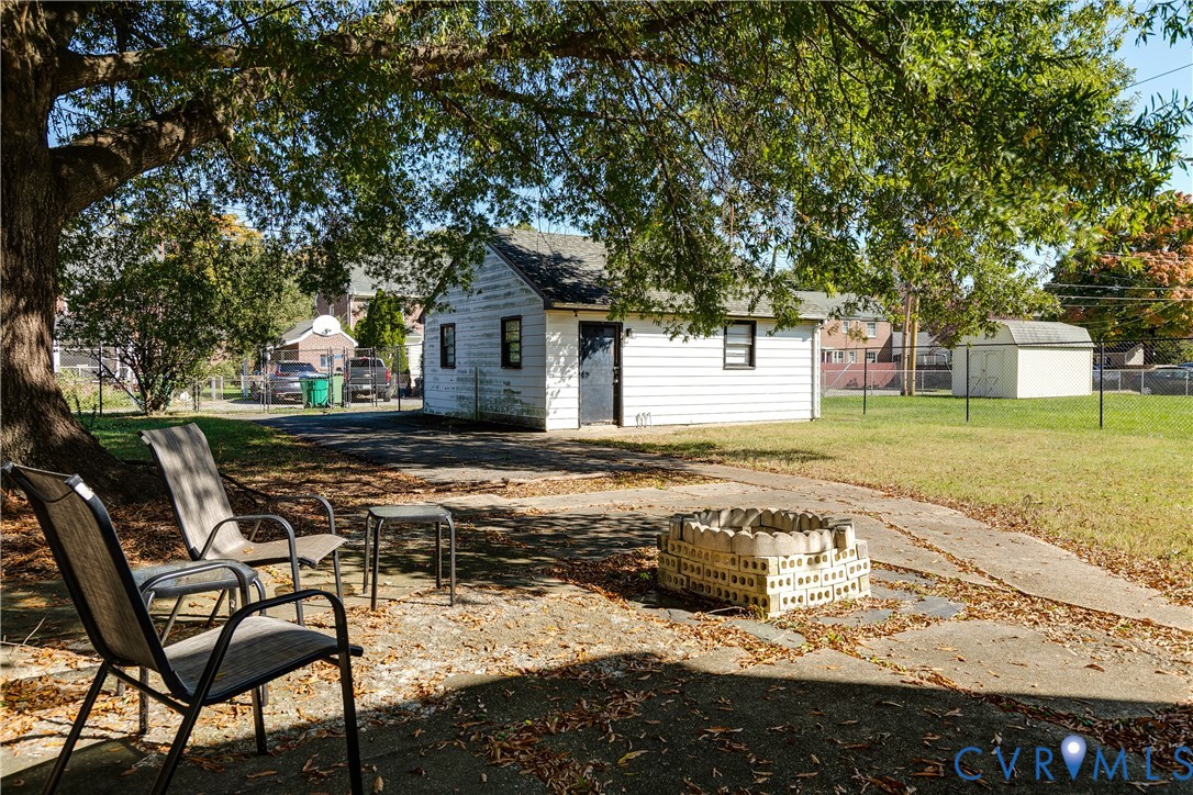 Concrete patio featuring a masonry fire pit, mature shade tree, detached structure with exterior siding, and fenced yard
