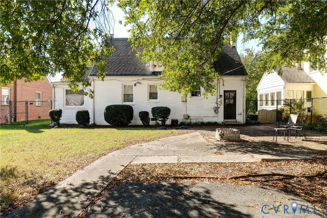 Exterior featuring a painted brick facade, steep-pitched gable roof, concrete patio, mature trees, and perimeter chain-link fencing