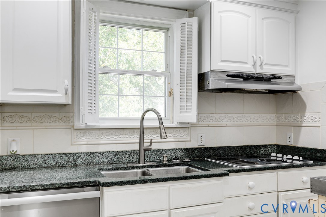 Kitchen featuring white cabinetry, stone-surface countertops, dual-basin stainless steel sink with high-arc faucet, integrated stainless steel range hood, and tile backsplash with decorative inset detailing