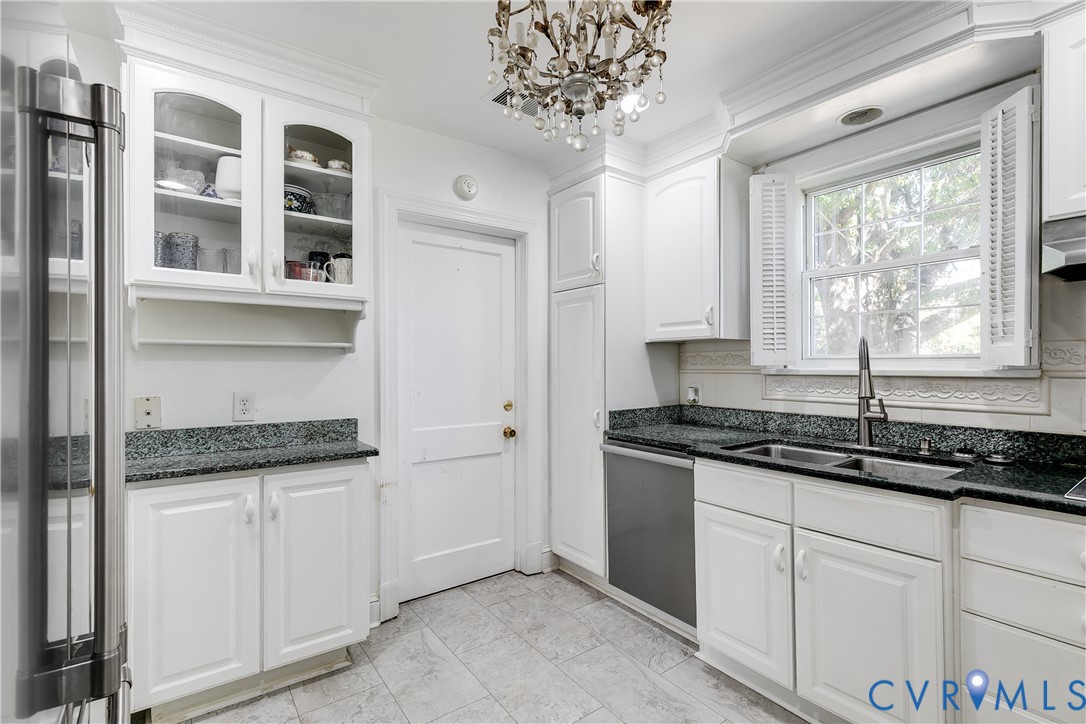 Kitchen featuring white cabinetry with glass-front display uppers, dark stone countertops, stainless steel undermount sink, ceramic tile flooring, and a tiered crystal chandelier