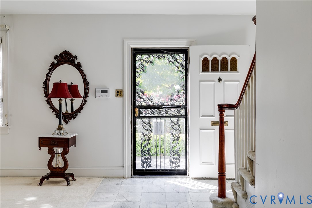 Entryway featuring a white paneled front door with decorative transom window, black metal screen door, stone-finish flooring, and a wooden staircase with a polished banister Entryway featuring a white paneled front door with decorative transom window, black metal screen door, stone-finish flooring, and a wooden staircase with a polished banister