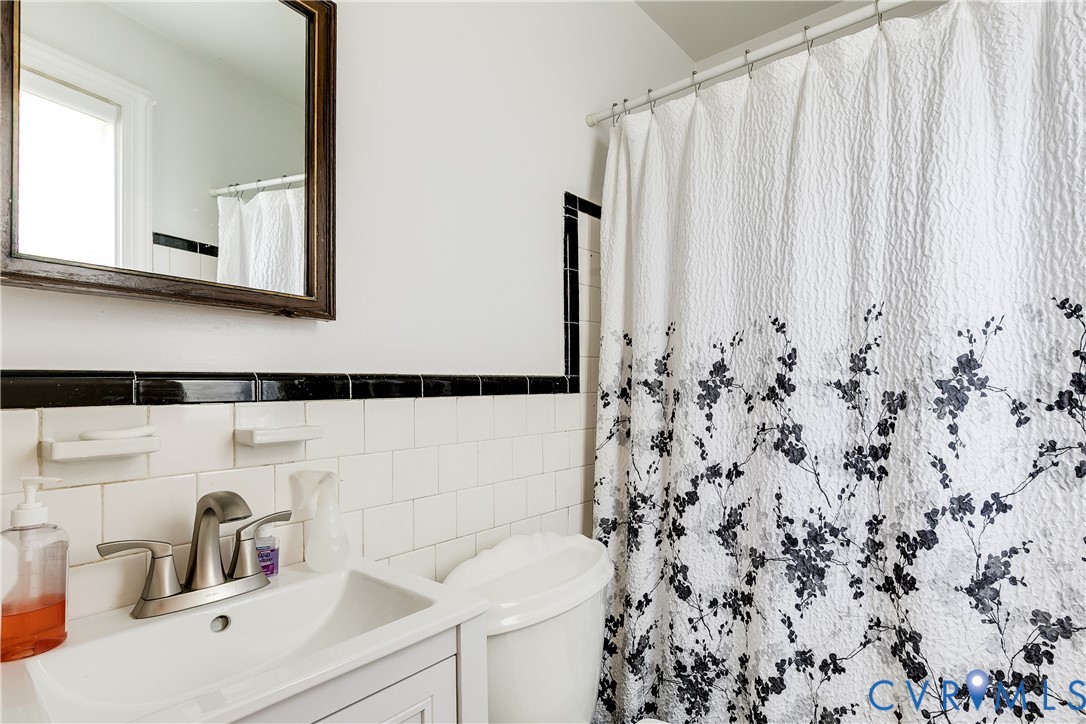 Wall-mounted mirror with a dark wood frame, white ceramic square wall tile, decorative black accent tile border, porcelain sink, and satin-finish metal faucet