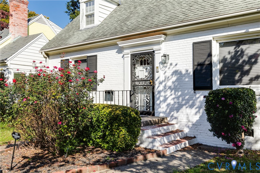 White brick exterior featuring dark shutters, a front dormer window, metal decorative storm door, and masonry steps White brick exterior featuring dark shutters, a front dormer window, metal decorative storm door, and masonry steps