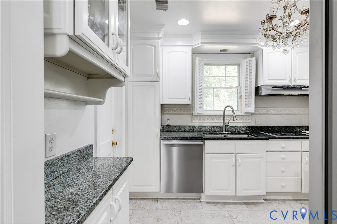 Kitchen featuring crisp white cabinetry, dark stone countertops, stainless steel dishwasher, decorative crystal chandelier, and tile flooring