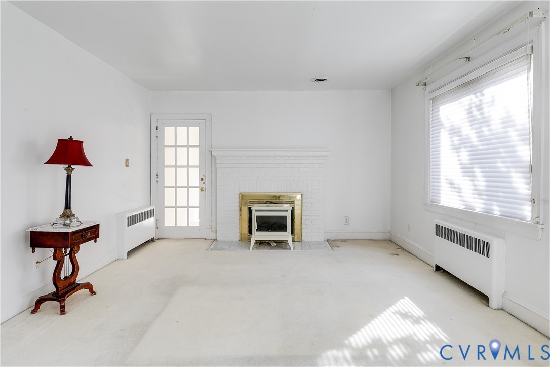 Interior space featuring a white painted brick fireplace with a metallic surround, a multi-lite glass panel door, and large windows with blinds Interior space featuring a white painted brick fireplace with a metallic surround, a multi-lite glass panel door, and large windows with blinds