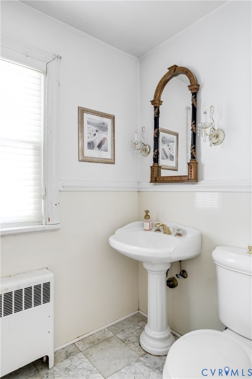 Pedestal sink with brass-finish hardware, wall-mounted arched mirror, dual crystal-style wall sconces, and natural stone tile flooring