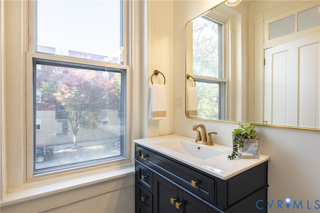 Dark-finish vanity featuring brass hardware and a white integrated sink, complemented by a gold-trimmed wall mirror, brass-toned faucet, and brass-finish towel ring