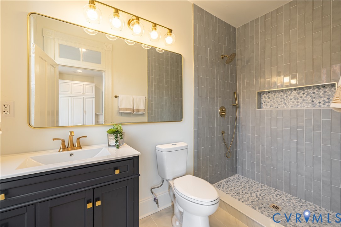 Dark-toned vanity with integrated white countertop, gold-finish faucet, wall-mounted light fixture, rectangular mirror, and walk-in shower with vertical tile surround and mosaic-tile niche