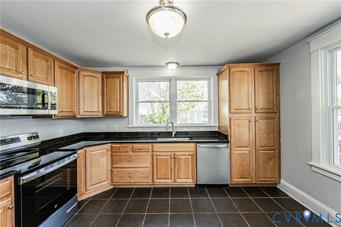 Kitchen featuring warm-toned cabinetry, dark stone countertops, stainless steel appliances, a central window above the sink, and tile flooring