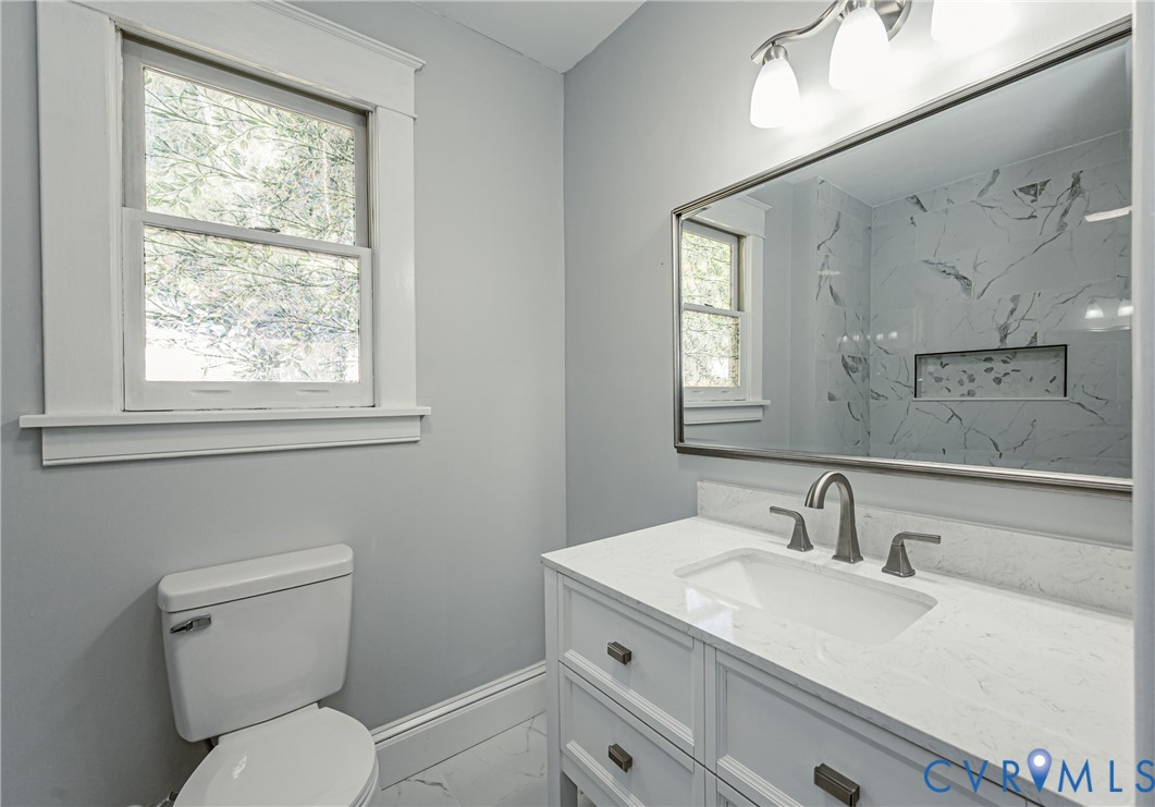 Vanity featuring a white countertop with an integrated basin, brushed metal hardware, wall-mounted mirror, and tile-surround shower with a built-in niche