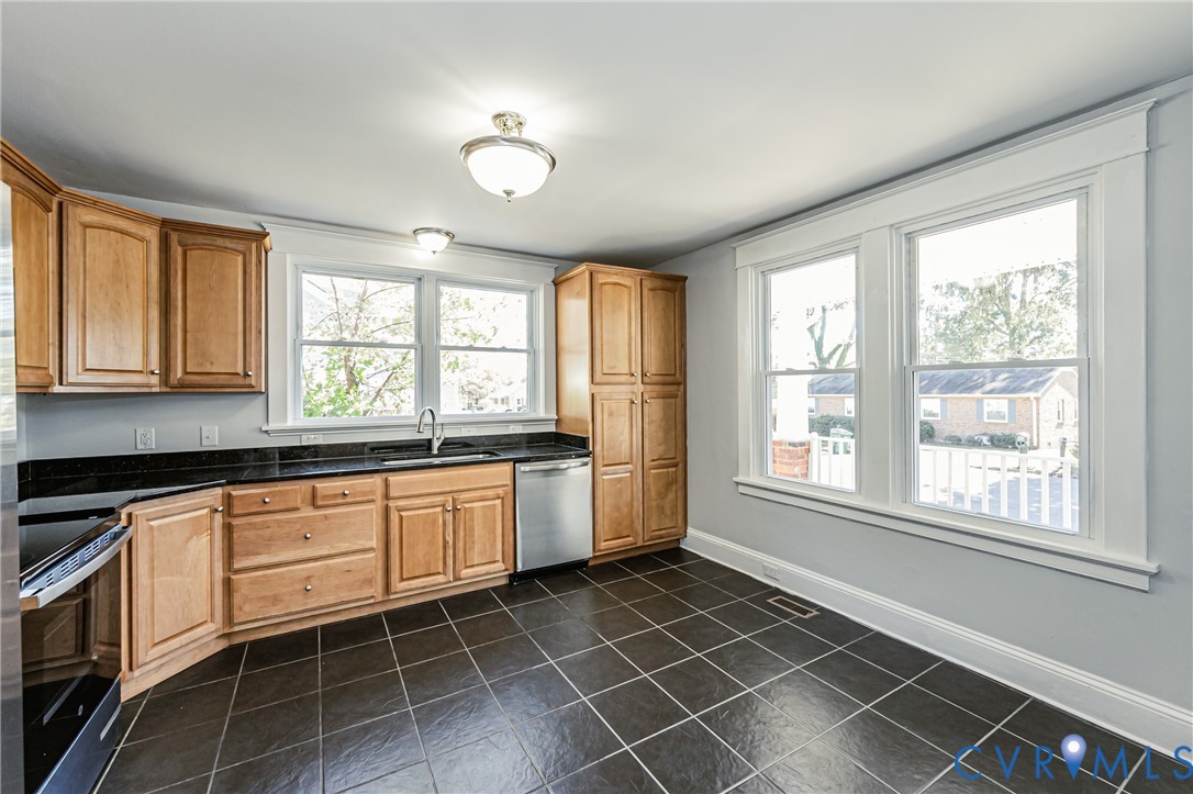 Kitchen featuring natural wood cabinetry, dark stone countertops, stainless steel appliances, large-format tile flooring, and dual multi-pane windows