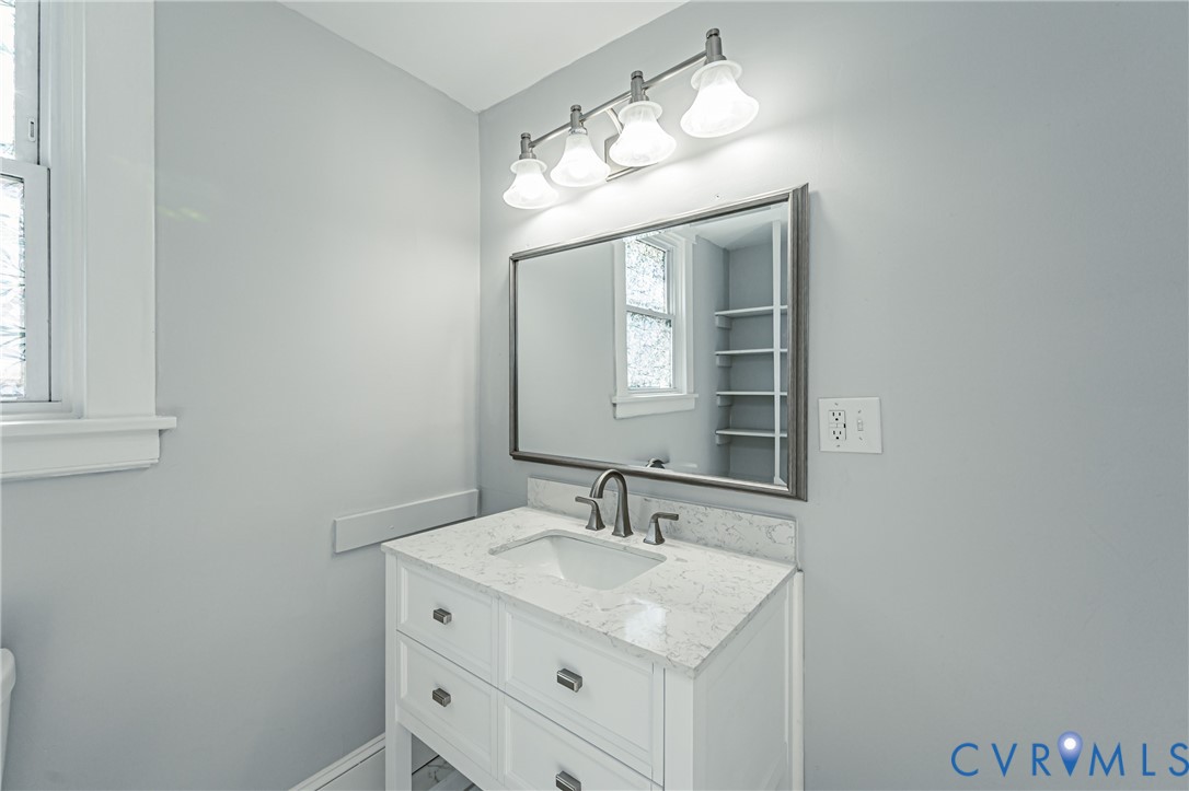 White vanity with a stone countertop, undermount sink, metal faucet, framed mirror, and four-light vanity fixture