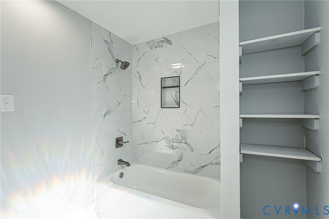 Bathtub with a shower head, wall-mounted faucet, built-in recessed wall niche, and stone-finish tile surround, complemented by a dedicated built-in shelving unit