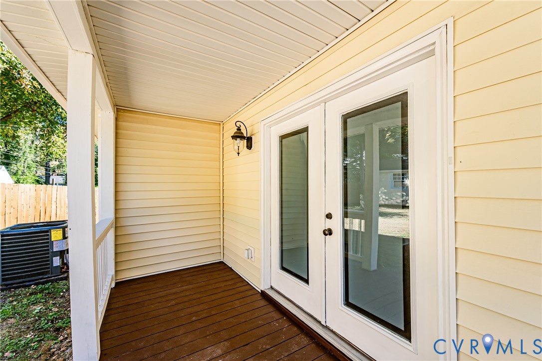 Covered porch with wood-finish decking, light-colored horizontal siding, dual glass-panel entry doors, and wall-mounted exterior light fixture