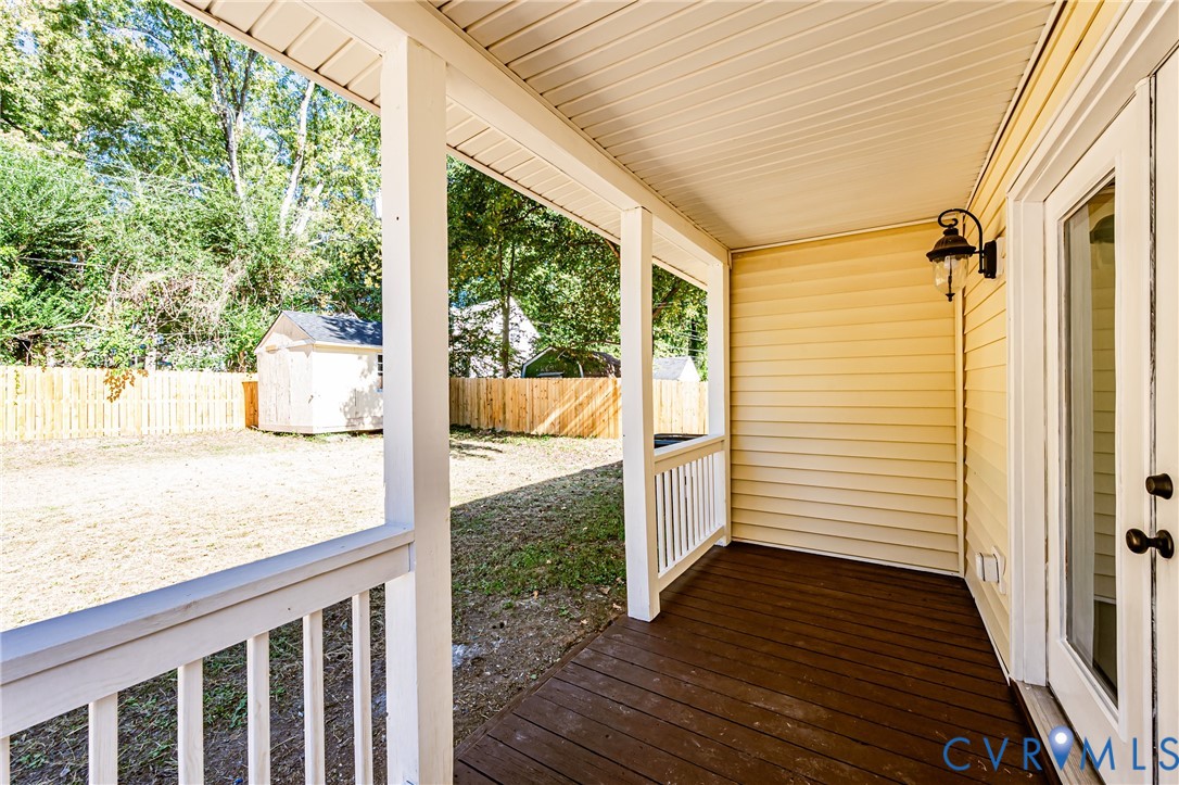 Covered porch with wood-finish decking, white baluster railing, wall-mounted exterior light fixture, and vinyl siding