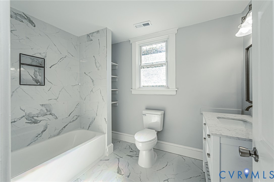 Bathroom featuring a vanity with a composite stone countertop, large-format tile flooring, a bathtub surround with floor-to-ceiling tile, built-in shelving, and a wall-mounted light fixture