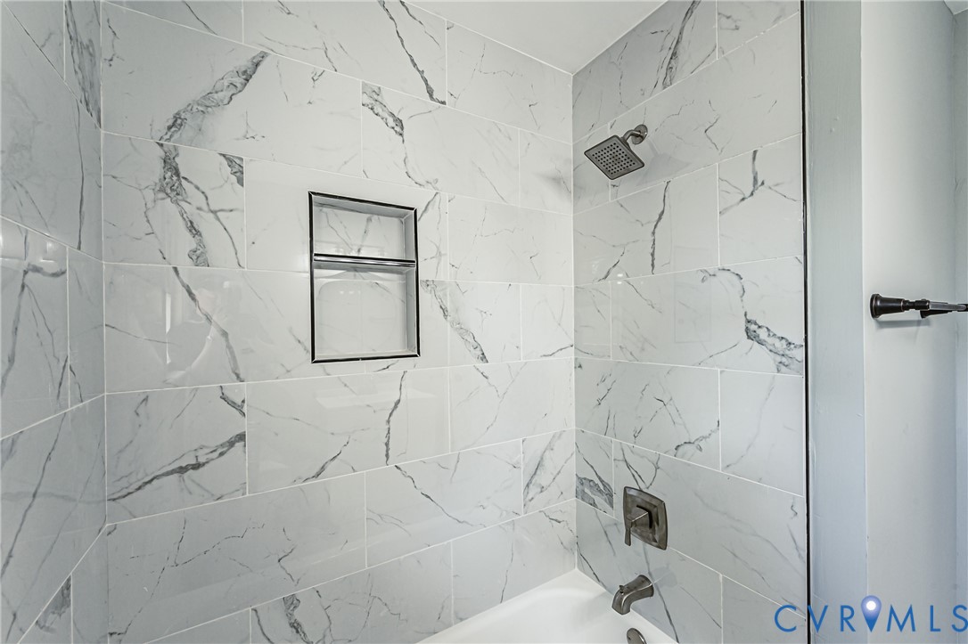 Bathtub surround featuring large-format white stone-patterned tile, integrated recessed shelving, a metallic shower head, and matching metallic faucet hardware
