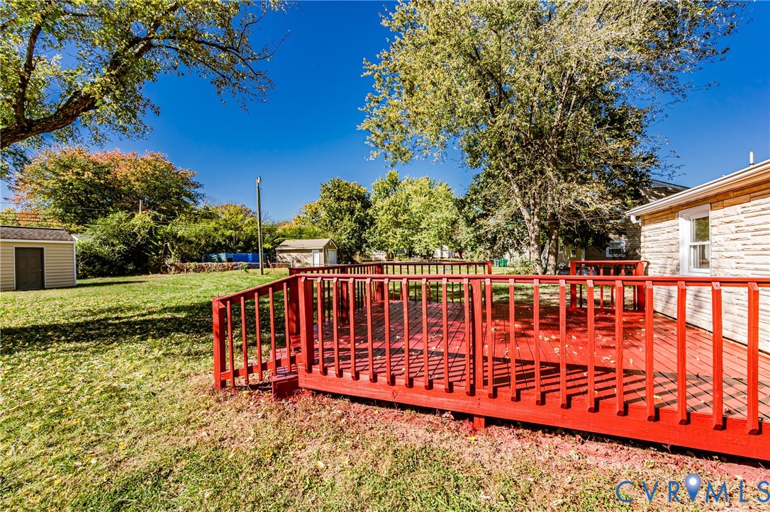 Backyard featuring a red-painted wood deck, expansive lawn, mature trees, and detached storage structure