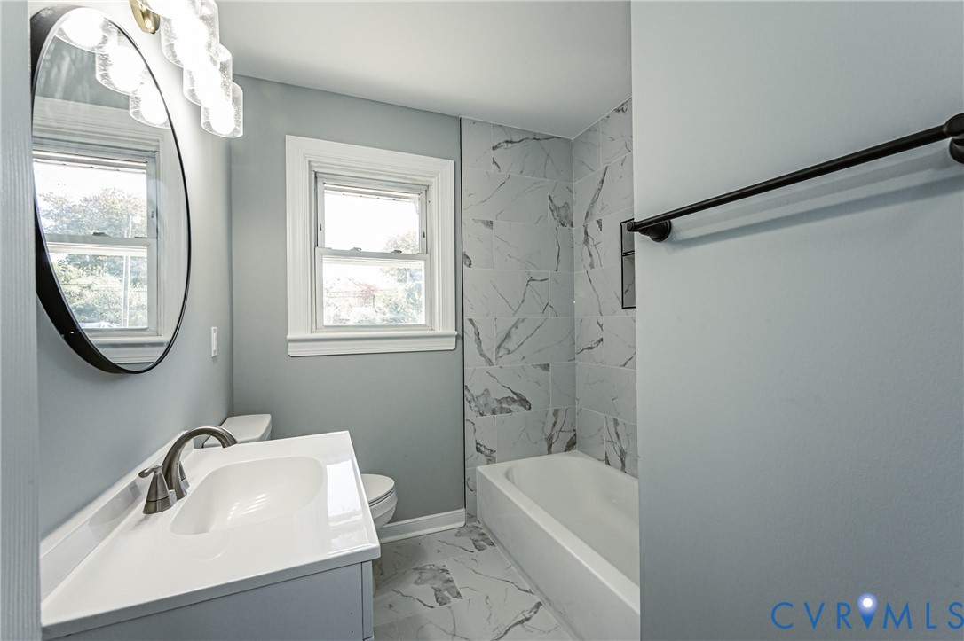 Bathroom featuring a vanity with an integrated sink and metallic faucet, a round wall-mounted mirror, overhead multi-globe lighting, a shower-tub combination with patterned wall tile, and coordinated floor tile