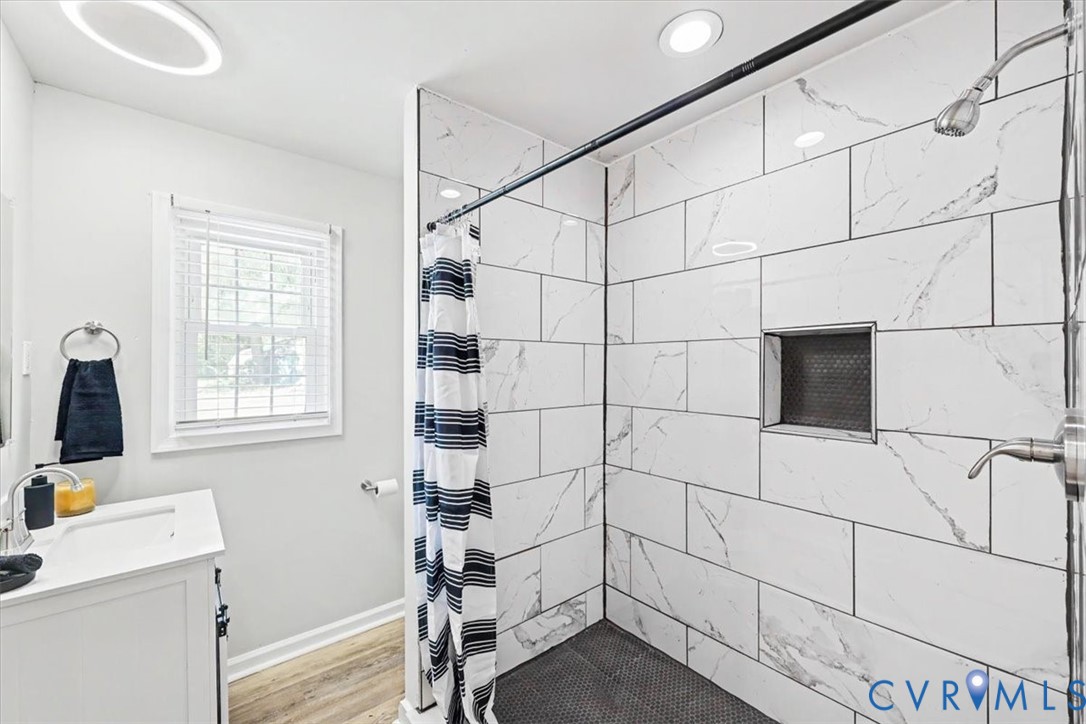 Bathroom featuring a shower stall with large-format stone-patterned wall tile, a recessed storage niche, wood-finish flooring, and a white vanity with an integrated basin